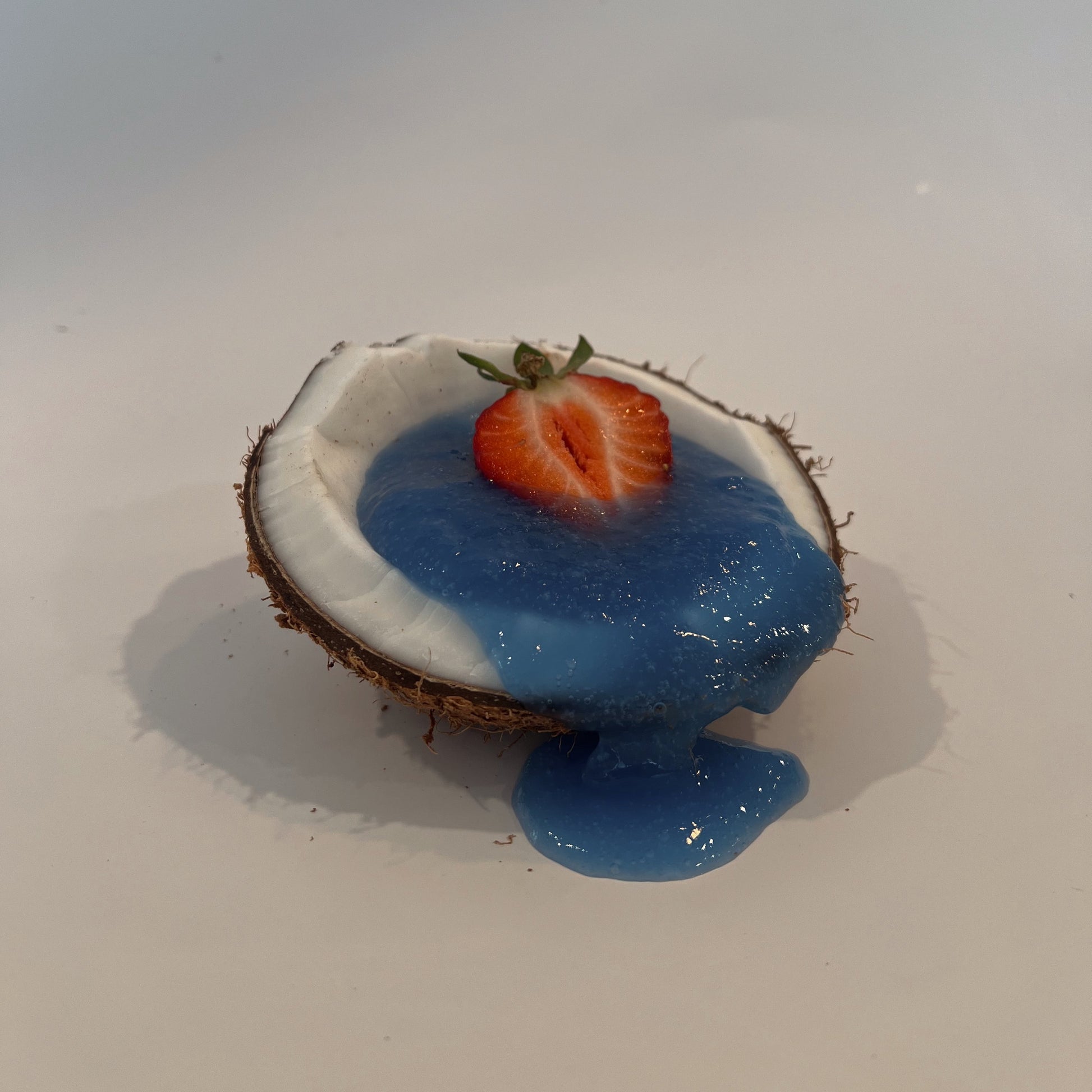 Blue Magik Sea Moss Gel from Organically Kismet poured into a halved coconut and topped with a sliced strawberry, with the vibrant blue gel flowing over the edge on a clean background.