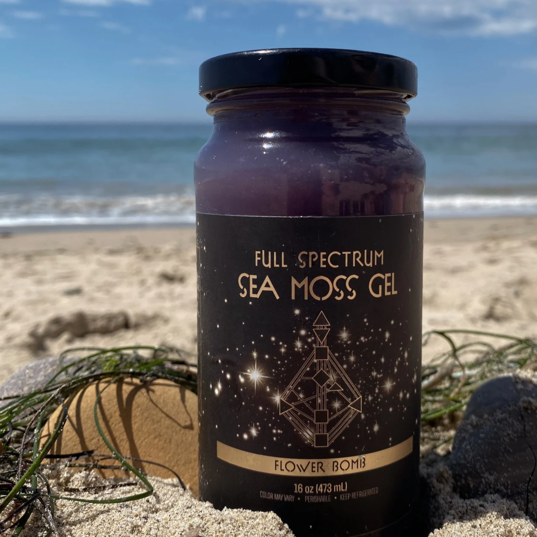 Jar of Organically Kismet Flower Bomb Full Spectrum Sea Moss Gel photographed on the beach with sand, seaweed, and the ocean in the background.