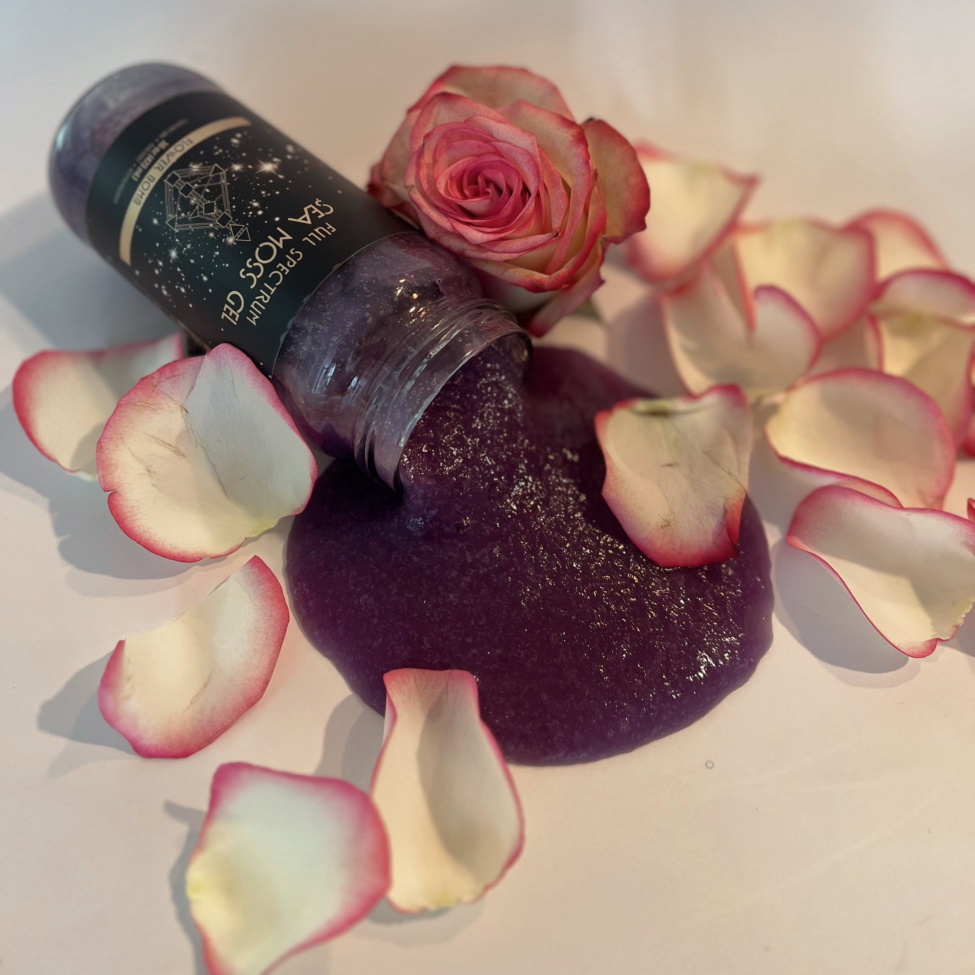 Organically Kismet Flower Bomb Sea Moss Gel spilling from a bottle surrounded by pink rose petals and a fresh rose, showcasing its rich purple color and luxurious texture.