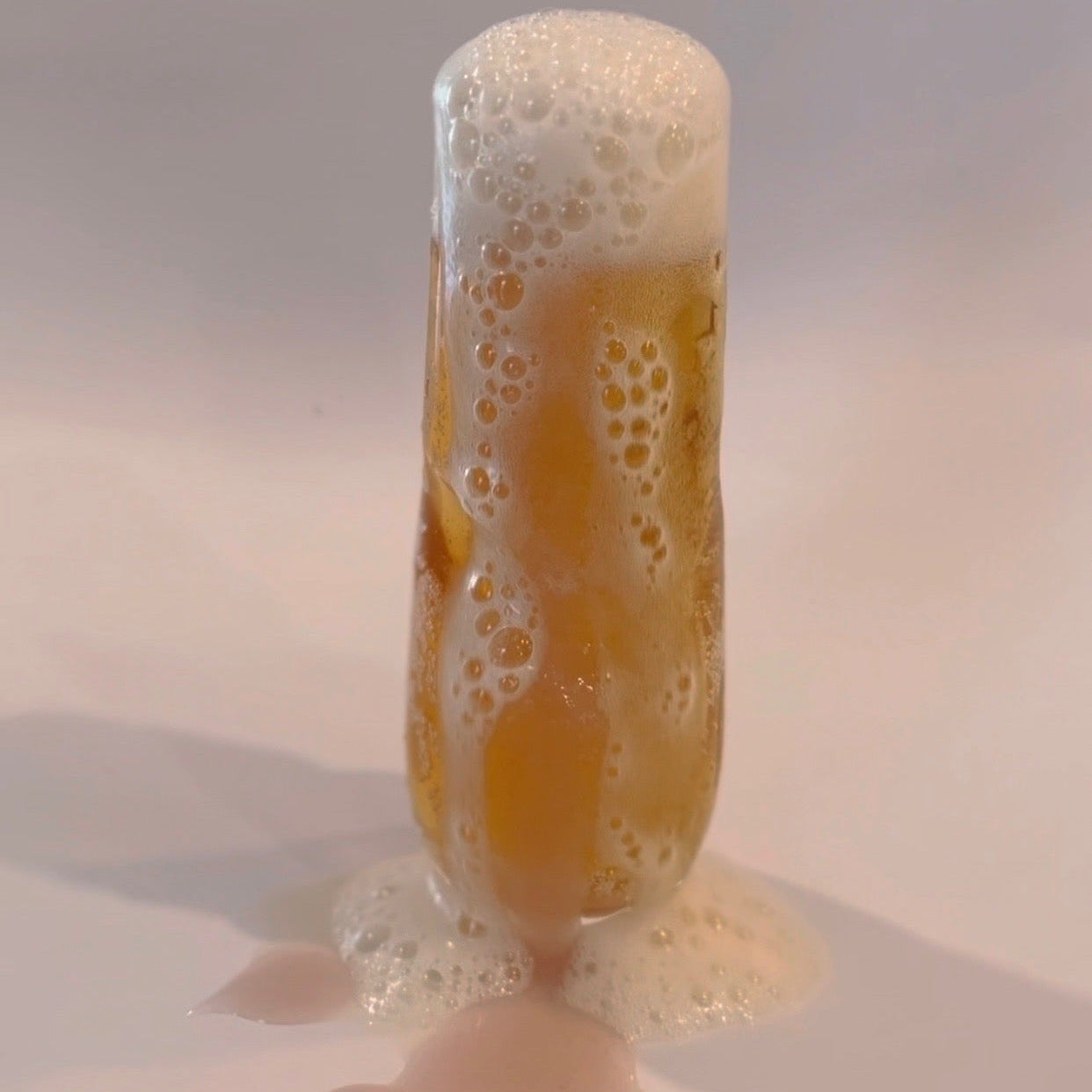 Glass of champagne mixed with Organically Kismet Original Sea Moss Gel, bubbling over with a foamy, golden effervescent overflow against a clean background.