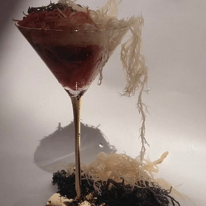 Raw full spectrum sea moss displayed in a martini glass with gold stem, overflowing with red, gold, and black sea moss strands on a reflective surface.